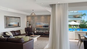 A modern living room with a large flat-screen TV, a brown leather sofa, a coffee table, and a view of a pool and outdoor area. - Apt 401 Luxury Serv Aparts-Estepona, 3 Bed, Heated 20m Pool, Jacuzzi - an Oasis (Estepona)