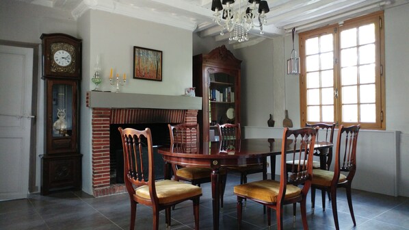 Comedor con chimenea de ladrillo, sillas de madera, una mesa redonda, un reloj de pie y un amplio ventanal de varios paneles.