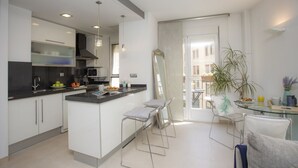A modern kitchen with white cabinets, a black countertop, and stainless steel appliances. There are two bar stools and a glass table with chairs. A large mirror and a potted plant are visible. - Singular Stays Poeta Querol (València)
