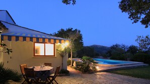 Eine Terrasse mit Tisch und Stühlen, ein Schwimmbecken und ein Gebäude mit gestreifter Markise.