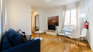 Una sala de estar moderna con un sofá azul, un televisor de pantalla plana, una zona de comedor con sillas blancas y un cuadro en la pared.