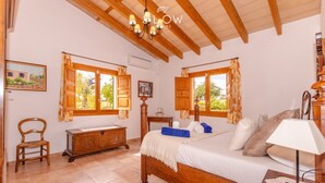 A bedroom with a wooden bed, two windows with curtains, a wooden chair, a bedside table, and a lamp.