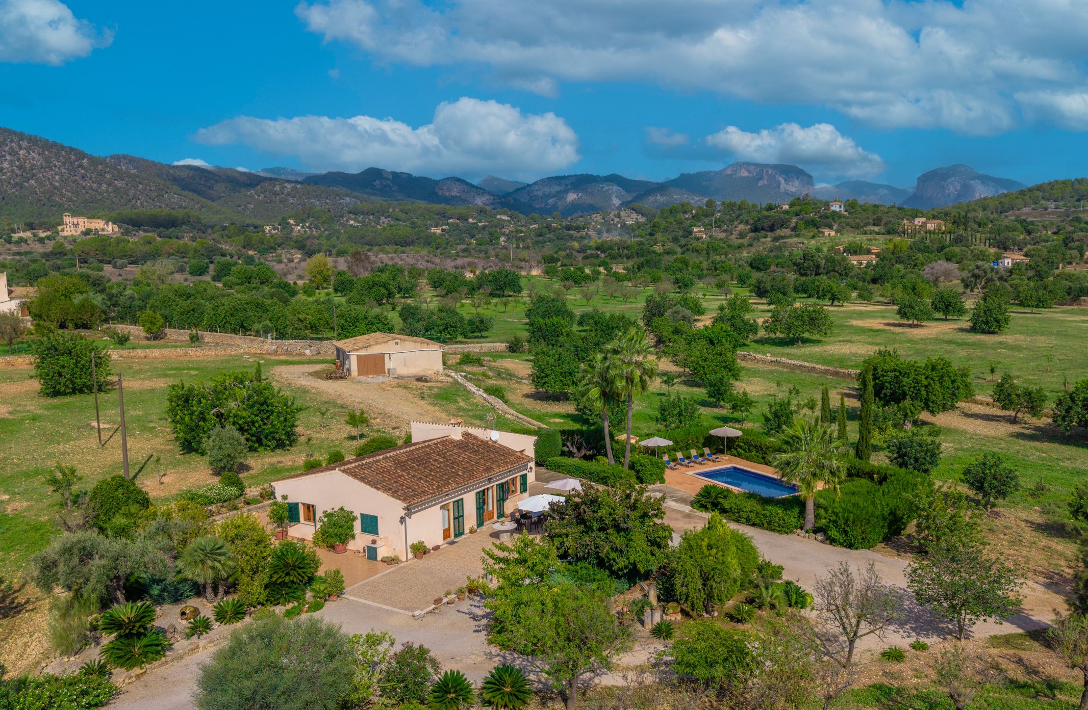 Paisaje rural con un edificio blanco de techo con tejas, piscina y jardín con diversos árboles y arbustos.