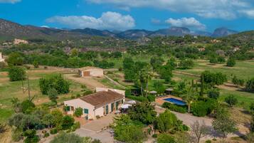 Paisaje rural con un edificio blanco de techo con tejas, piscina y jardín con diversos árboles y arbustos.