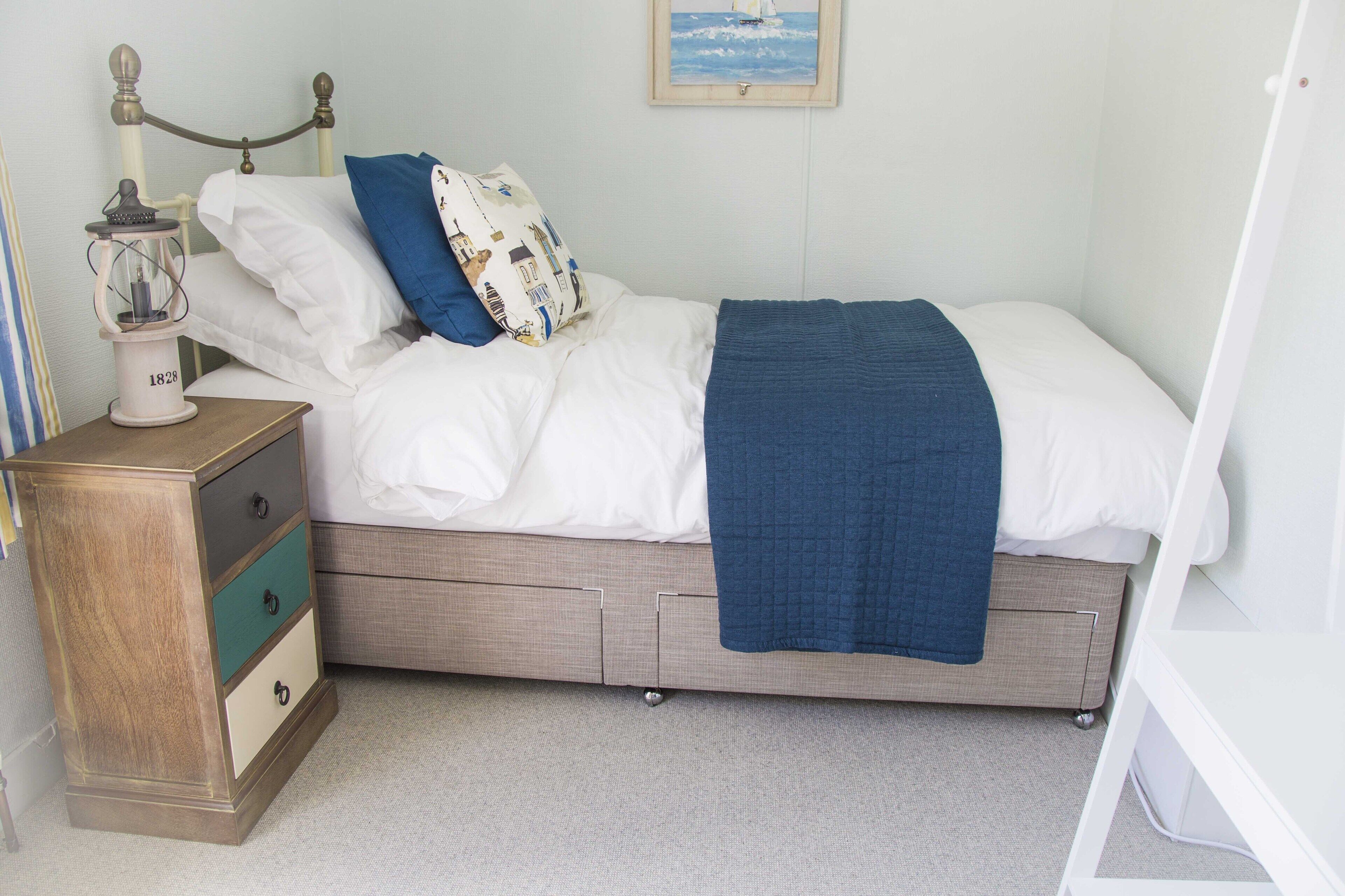 Een net opgemaakt bed met een blauwe deken, een nachtkastje met een lamp en een aan de muur hangend schilderij.