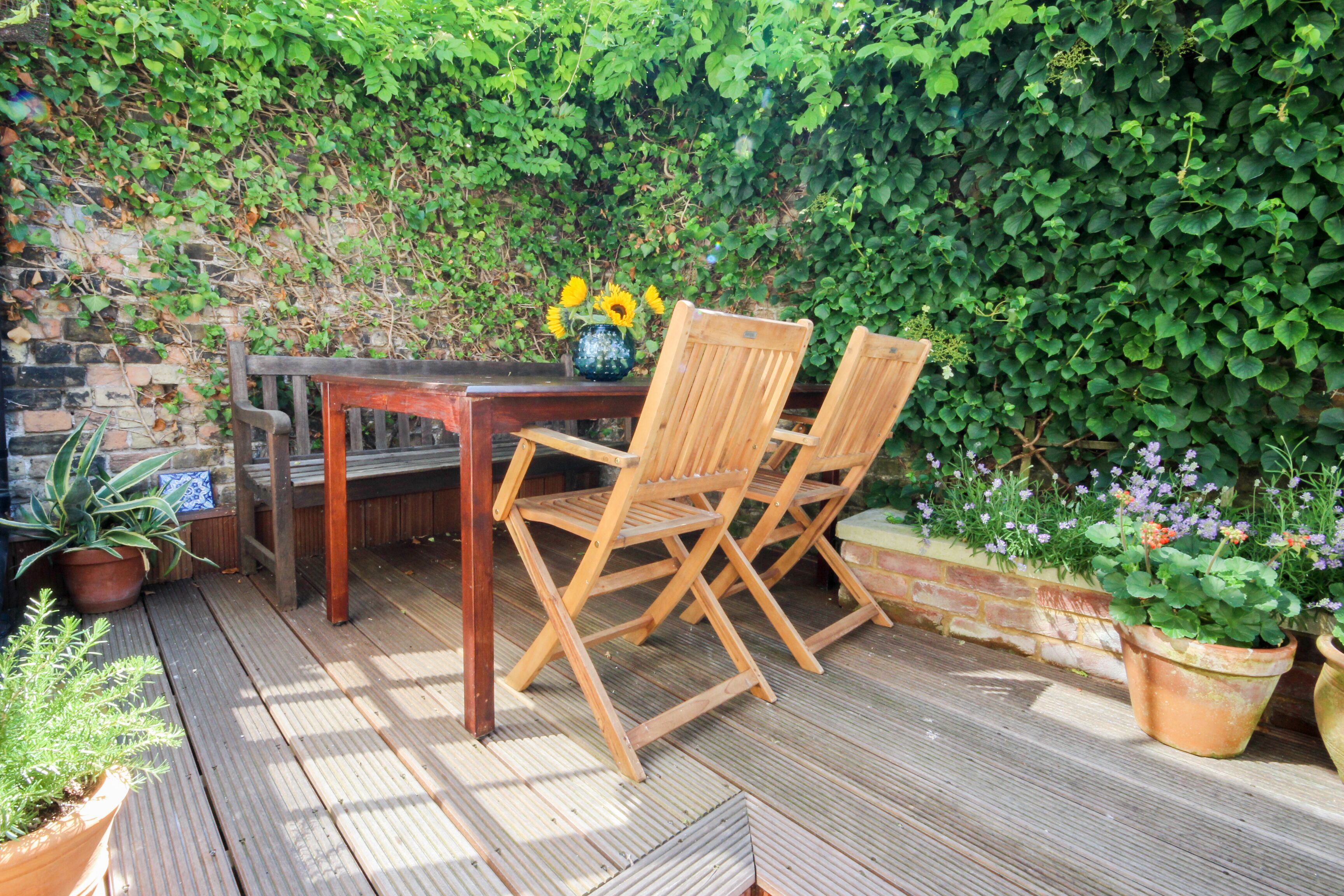 Una terraza de madera con una mesa y sillas, rodeada de plantas en macetas y un muro de ladrillo.
