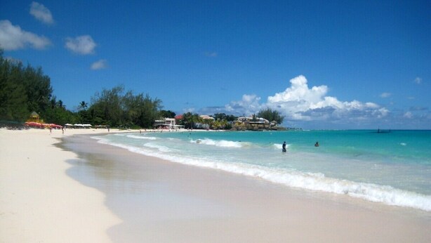 A beach with clear turquoise water, a sandy shore, and a few buildings in the background.