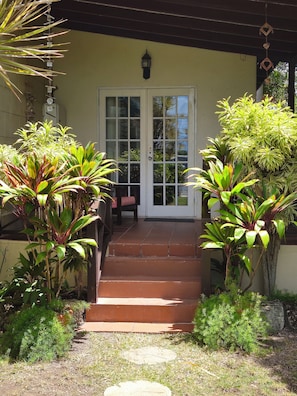 Une véranda couverte, avec un escalier menant à une porte, le tout entouré de différentes plantes.