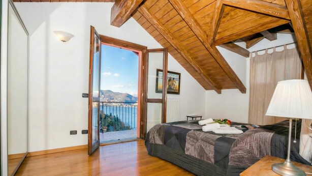 A bedroom with a bed, a nightstand, a lamp, a picture on the wall, and a view of the outdoors.