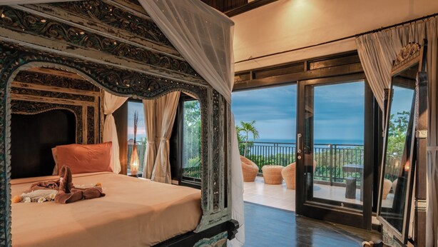 A luxurious bedroom with a large bed, ornate canopy, and a balcony with a view of the ocean.