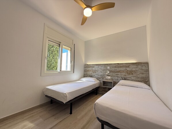 A room with two single beds, a ceiling fan, a window with a view of greenery, and a wall-mounted nightstand.