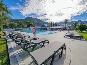 A poolside area with lounge chairs and a lifebuoy, surrounded by greenery and buildings, with mountains in the background. - Agi Rescator 330 Apartamento for rent in Roses Girona. (Rosas / Roses)