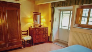 A bedroom with a large wooden wardrobe, a dresser, a chair, a bed with a blue bedspread, and a window with curtains.