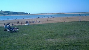 Une plage avec des gens et une zone herbeuse avec une chaise et un parasol.