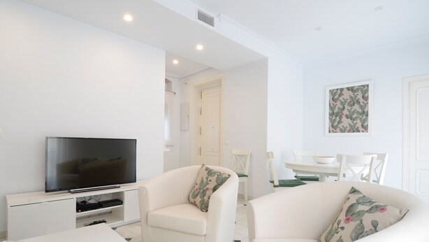 A modern living room with a flat-screen TV, two white armchairs with floral cushions, a white coffee table, and a dining area with a white table and chairs. - Among magnolias, in the heart of the city ... (Córdoba)