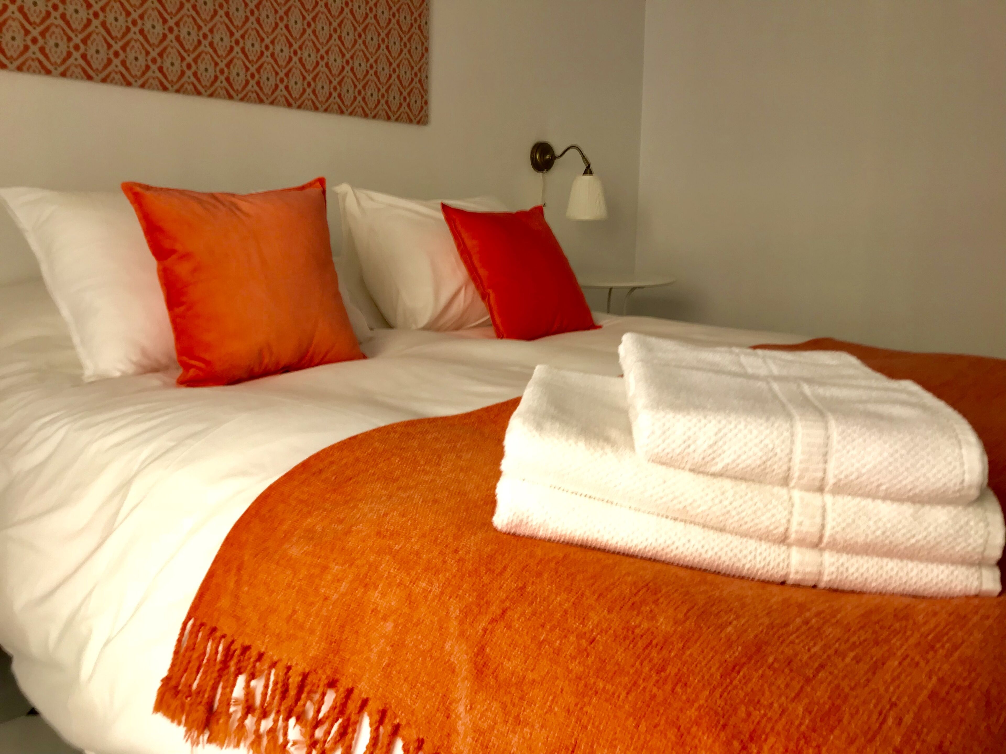 Una cama bien hecha con ropa de cama blanca y naranja, una lámpara blanca en la mesita de noche y una pared con diseño.