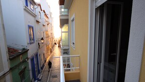 A narrow street with buildings on both sides, a staircase leading to a balcony, and a person walking down the street.