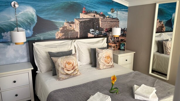A hotel room with a large mural of a coastal scene, a bed with a patterned bedspread, two bedside tables with lamps, and a mirror.