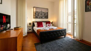 Ein Hotelzimmer mit einem schwarzen möblierten Bett, einem großen Fenster mit Vorhängen, einem wandmontierten Fernseher und einem Stadtbild in einem Rahmen.
