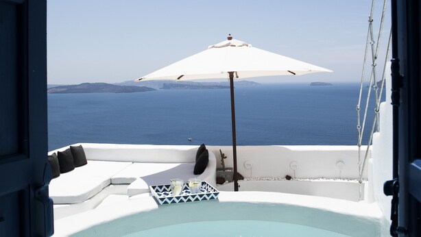 An open doorway to a balcony with a hot tub, a white umbrella, and a view of the sea.