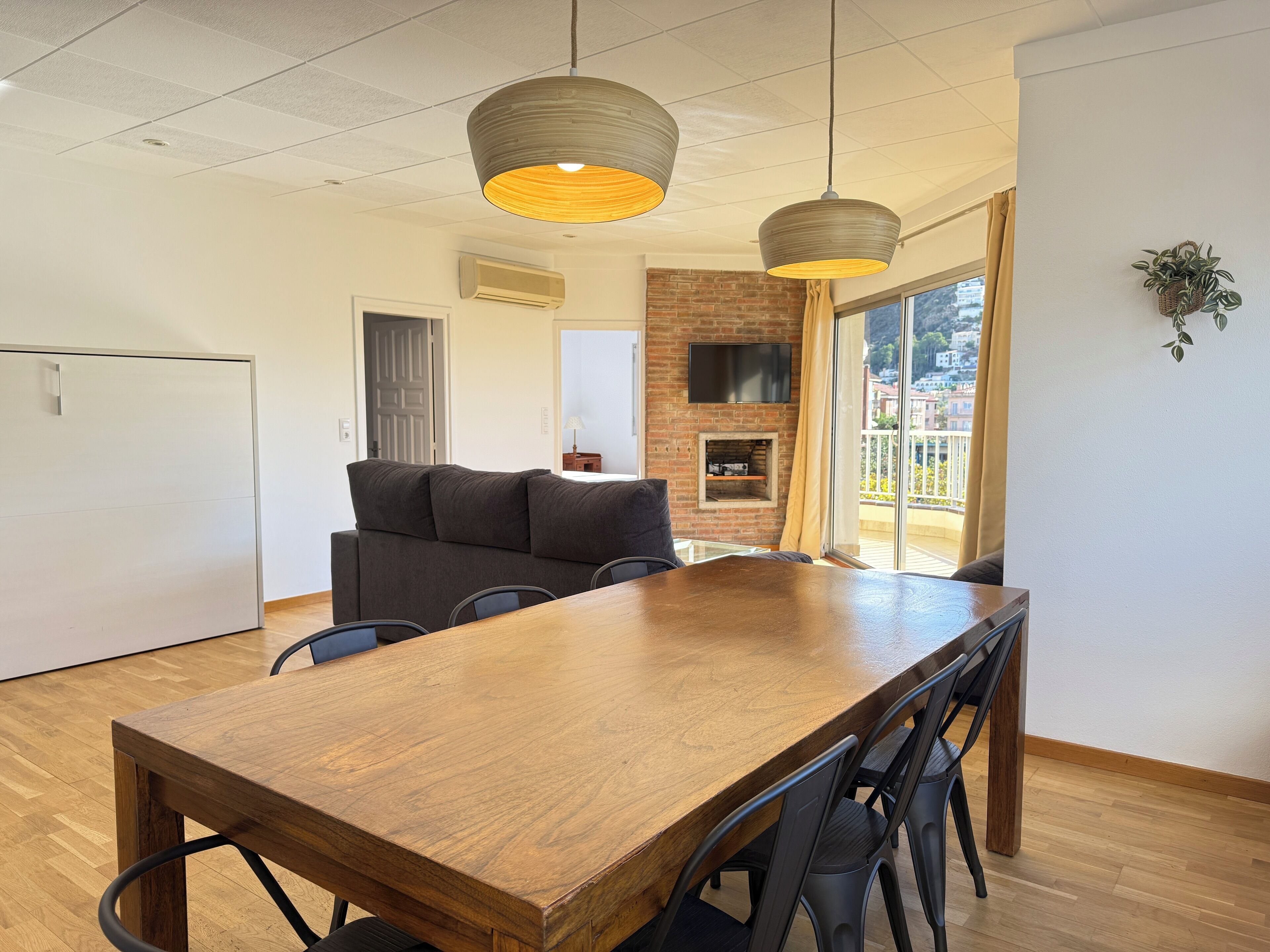 A modern dining area with a wooden table and chairs, a sofa, and a TV mounted on a brick wall.