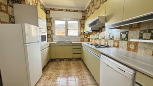 A compact kitchen with a refrigerator, sink, stove, and dishwasher. The walls have a patterned tile design, and there is a window above the sink. - Agi Peater 3A Apartamento for rent in Roses Girona. (Roses)