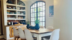 Een eetruimte met een houten tafel en witte stoelen, een keuken met planken en een raam, en een hanglamp.