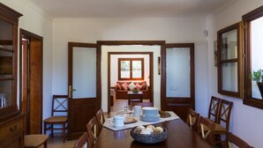 A dining room with a large wooden table set for six, flanked by wooden chairs. A cozy seating area with a sofa and cushions is visible in the background. - Cottage `Es Pinaró` (Manacor)
