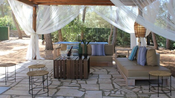 An outdoor patio area with a wooden pergola, cushioned seating, a wooden table with a bottle and glasses, and a hanging light fixture. - Cottage `Es Pinaró` (Manacor)