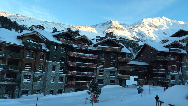 Un chalet de montagne enneigé, doté de plusieurs balcons, avec une piste de ski en arrière-plan.
