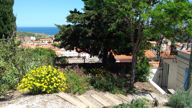 Un jardín con terraza de madera, diversas plantas y vistas a un pueblo costero con edificios y el mar.