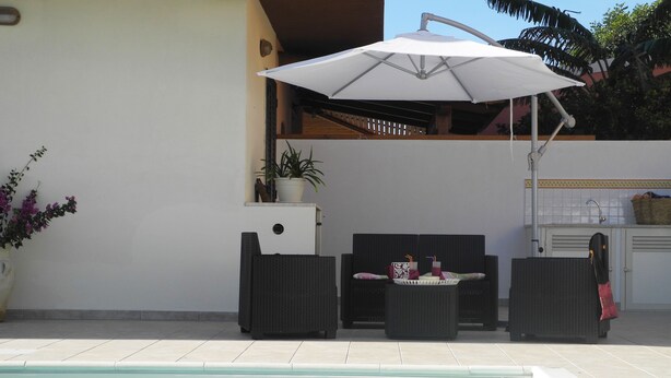 A poolside area with a white umbrella, two wicker chairs, a small table with drinks, and a potted plant.