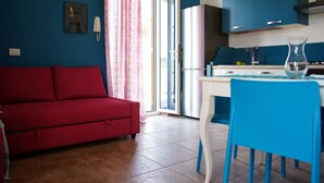 A room with a red sofa, a blue chair, a kitchen with blue cabinets, and a glass vase on the table.