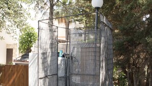 Un área pequeña y cerrada para ducha al aire libre, con valla metálica, manguera de agua y un accesorio de iluminación.