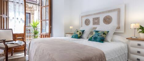 A bedroom with a large bed, a wooden chair, a nightstand, and a window with curtains.