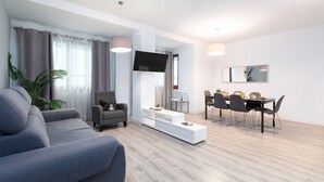 A modern living room with a grey sofa, a TV mounted on the wall, a dining area with a table and chairs, and a large potted plant.