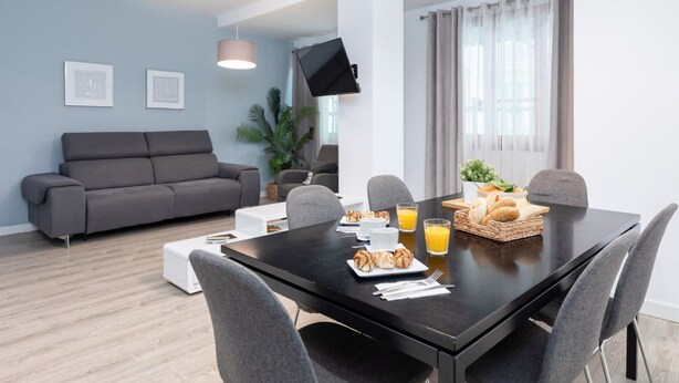 A modern living room with a grey sofa, a dining table with breakfast items, and a flat-screen TV mounted on the wall.