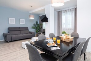 A modern living room with a grey sofa, a dining table with breakfast items, and a flat-screen TV mounted on the wall. - A (VA012.2) TH Ayuntamiento 1B (Valencia / València)