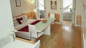 Une chambre confortable avec un lit, une table de chevet et un petit bureau. On y trouve une fenêtre avec des rideaux, un climatiseur fixé au mur et une porte donnant sur une autre pièce.