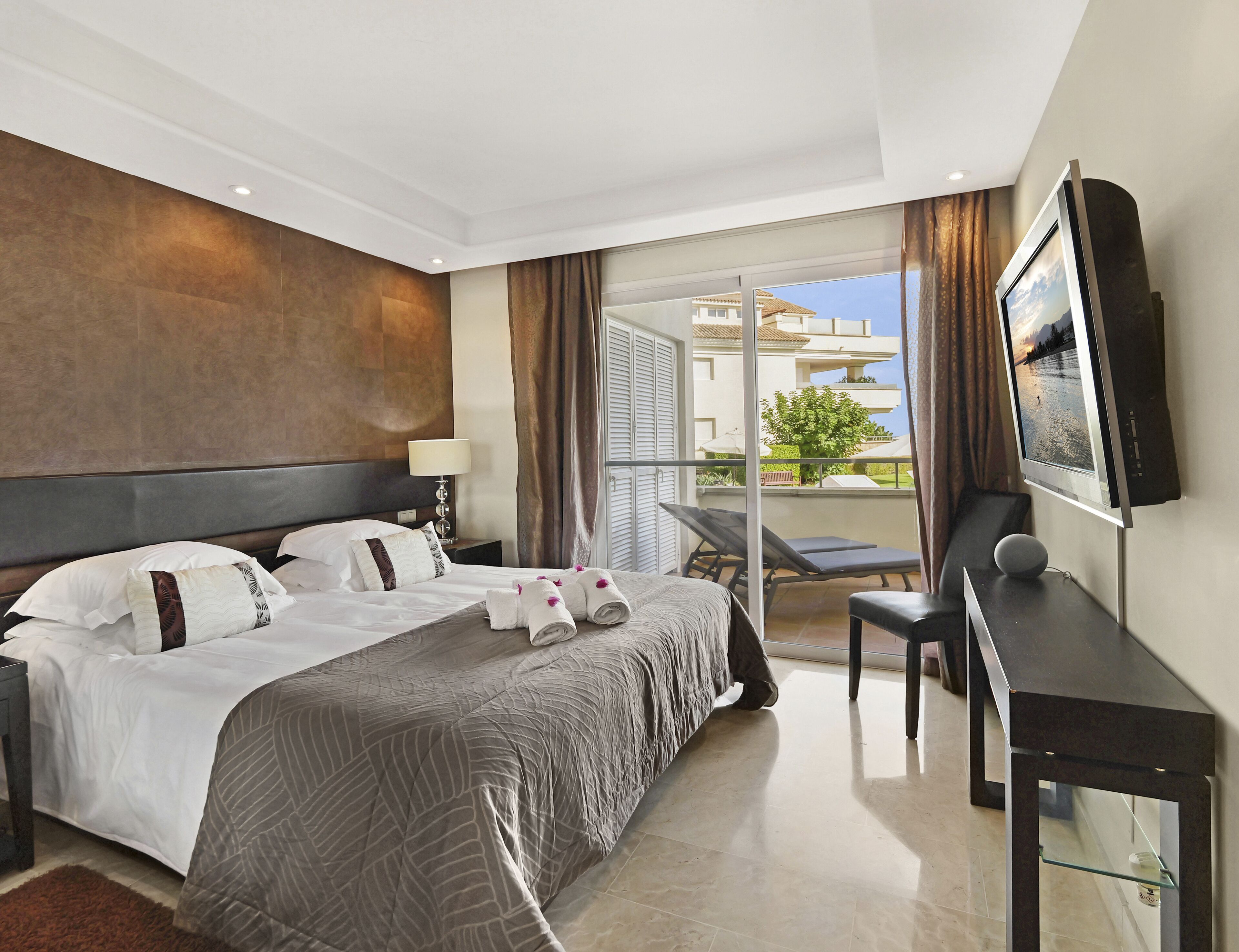 A hotel room with a large bed, a desk with a flat-screen TV, a chair, and a balcony with a view of buildings and trees.