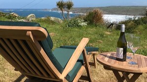 Una silla y mesa de madera con una botella y vasos sobre una colina cubierta de hierba con vistas al océano.