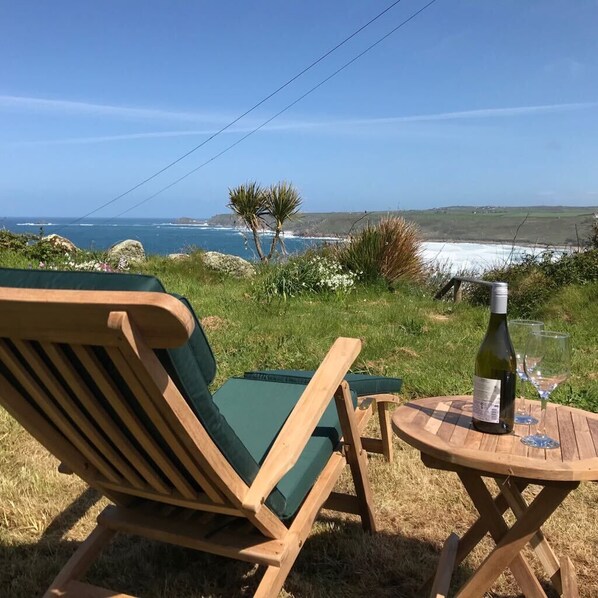 A wooden chair and table with a bottle and glasses on a grassy hill overlooking the ocean.