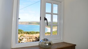 Una ventana con vista a un paisaje costero, un muro de piedra y una maceta de metal sobre un repisa de madera.