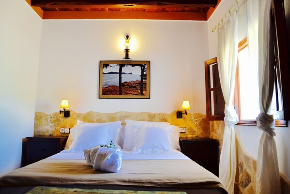 Una cama bien hecha con ropa blanca, dos lámparas de noche, un cuadro en la pared y una ventana con cortinas.