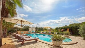 Zona de piscina con sillones reclinables y sombrillas, rodeada de vegetación y un cielo despejado.