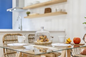 Une table de petit-déjeuner avec un plateau en verre, des chaises en osier, un panier de viennoiseries, une tasse blanche, un fruit jaune et une pomme.