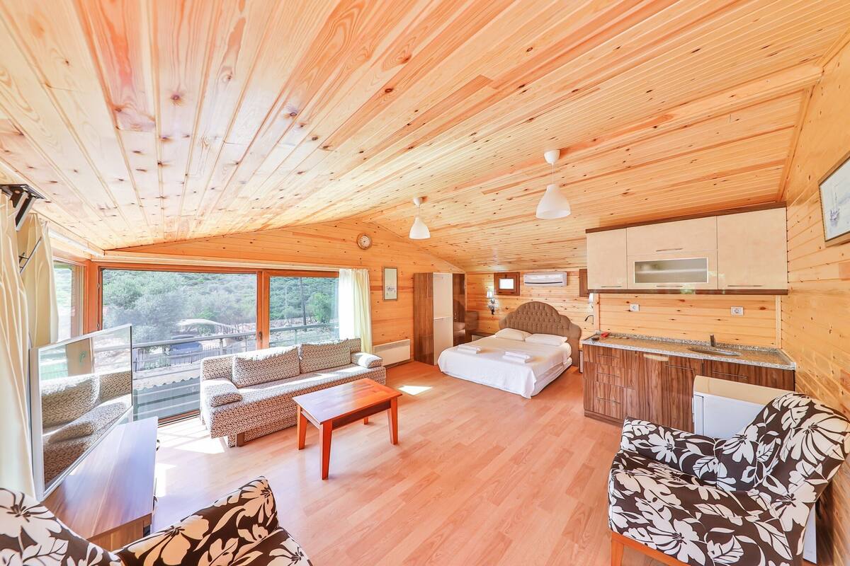 Interior de una cabaña de madera con sofá, cama, zona de comedor y vistas amplias por las ventanas.