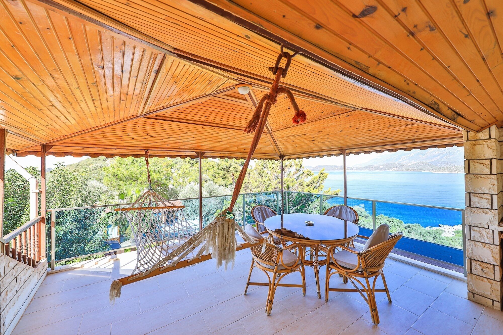 A covered outdoor seating area with a table and chairs, overlooking a scenic view of a body of water and greenery.