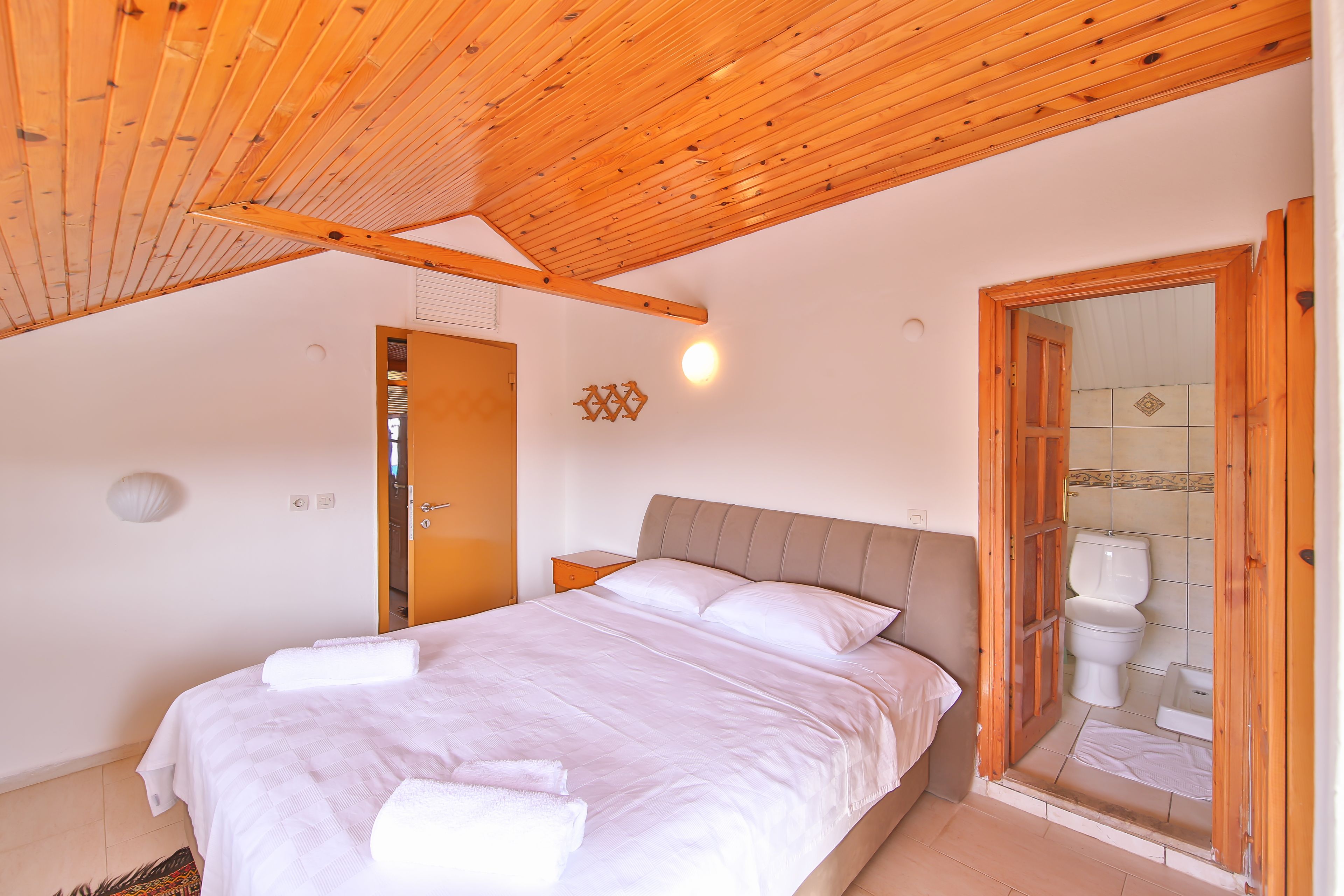 A bedroom with a wooden ceiling, a bed with white linens, a nightstand, and a bathroom with a toilet and shower.