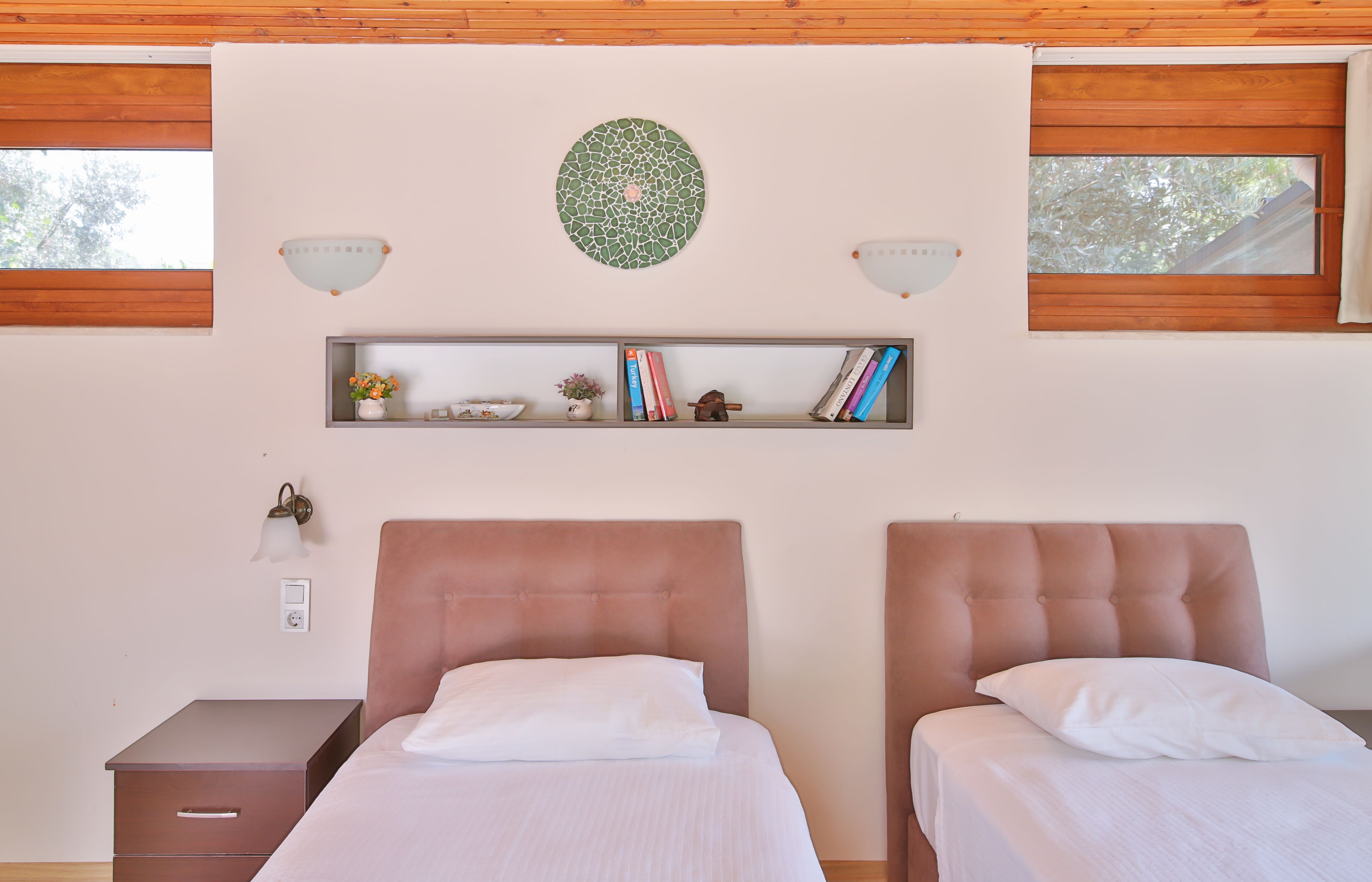 Quarto com duas camas, uma estante com livros e objetos decorativos, e um aplique na parede.